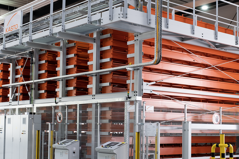 Großes mehrstöckiges orangefarbenes Lagerregalsystem mit Metallgitter-Sicherheitsabsperrung und zwei Bedienpulten im Vordergrund in einem Industriegebäude