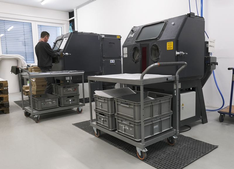 Zwei Sandstrahlkabinen mit Handschuheinsätzen in einem Werkstattbetrieb, davor Metallwagen mit grauen Kunststoffboxen und ein Mann, der eine der Kabinen bedient.