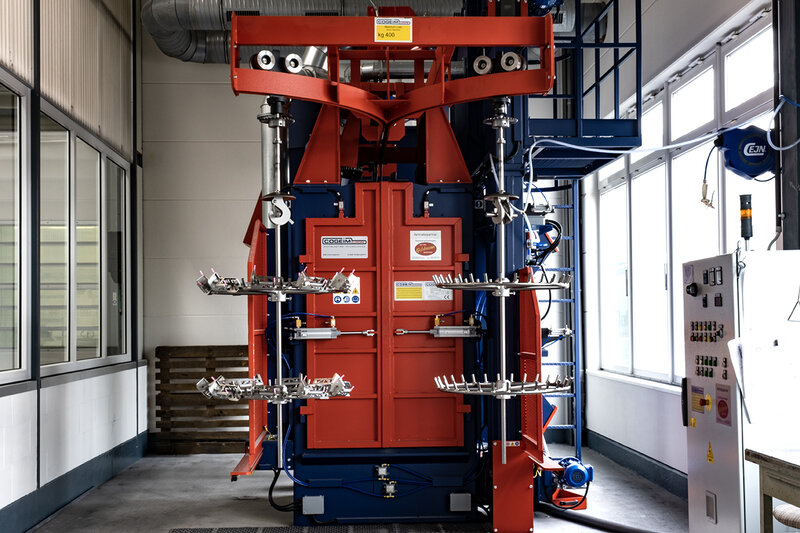 Industriemaschine mit roten und blauen Teilen, mehreren rotierenden Halterungen und Haken in einer hellen Fabrikumgebung mit Kontrollpanel und großen Fenstern