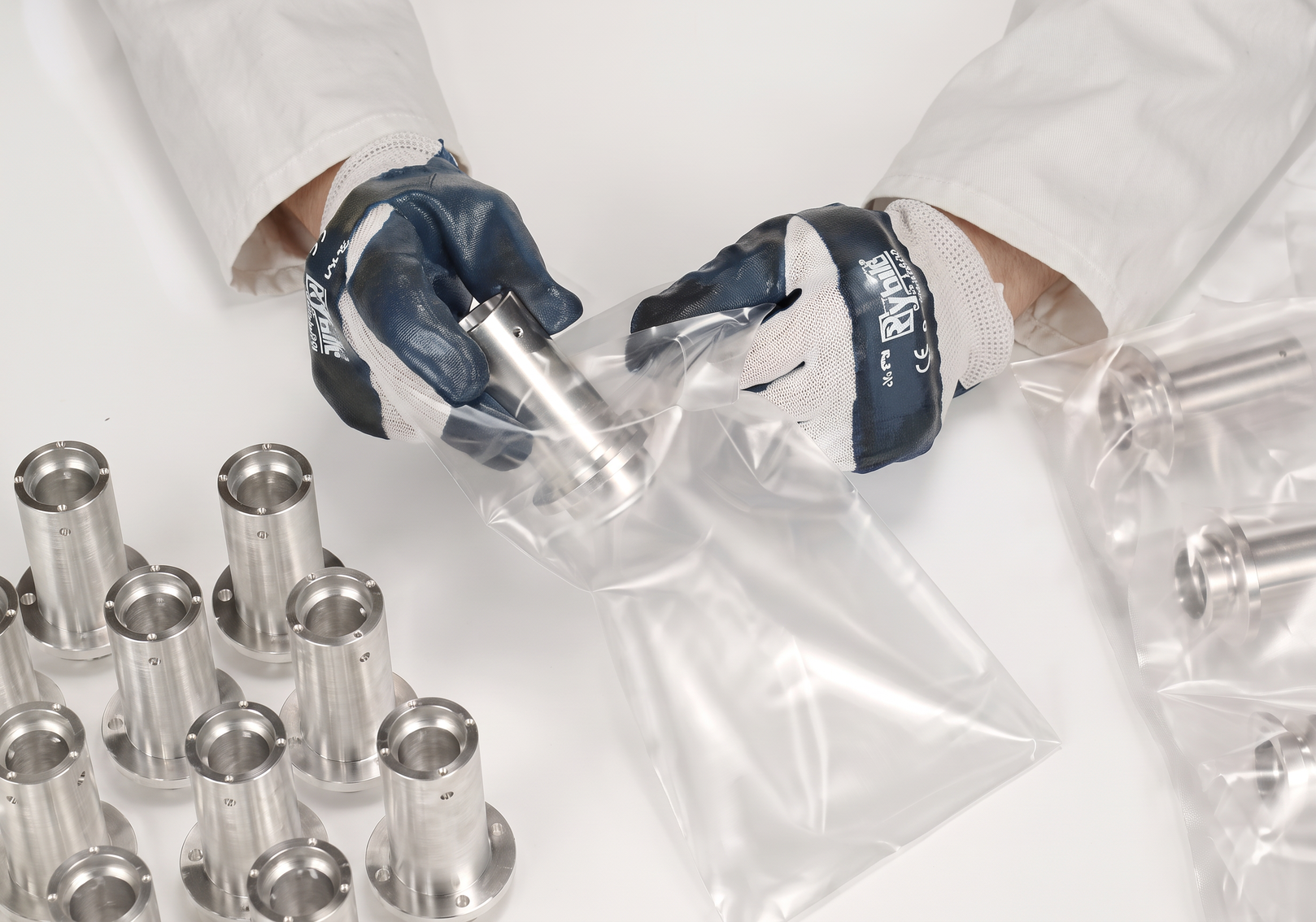 Mitarbeiter verpackt Aluminium-Bauteil in transparenten Kunststoffbeutel, umgeben von weiteren Bauteilen auf weißem Untergrund
