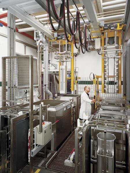 Industrielle Anlage mit mehreren Behältern und Hebevorrichtung, ein Mann im weißen Laborkittel bedient die Maschine zum Reinbeizen von Aluminiumbauteilen