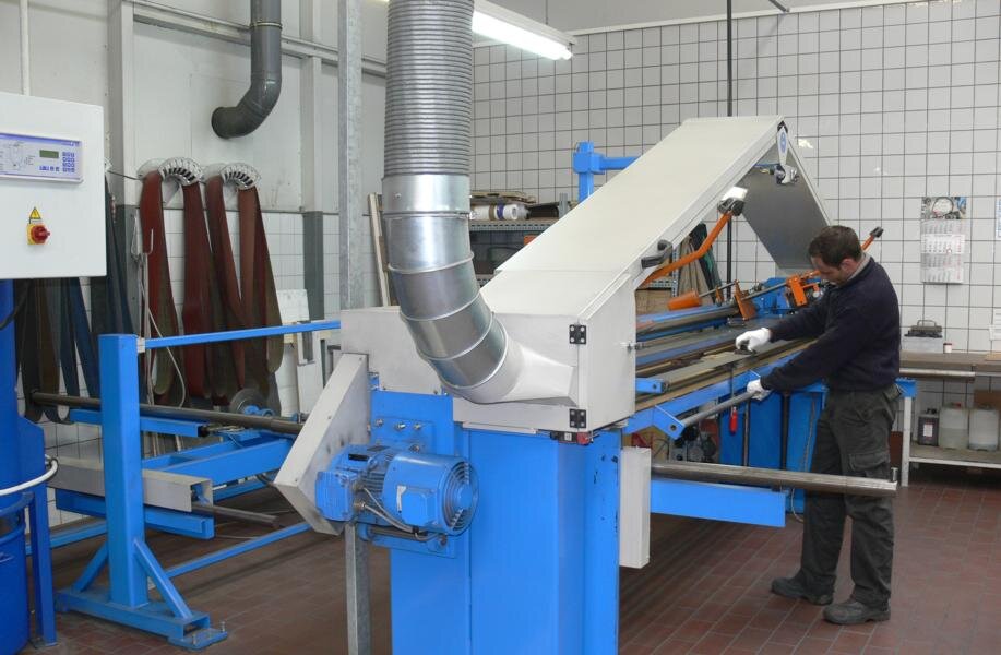 Ein Mann bedient eine blaue und weiße Doppelbandschleifmaschine in einer Werkstatt mit Fliesenwänden und rotem Boden