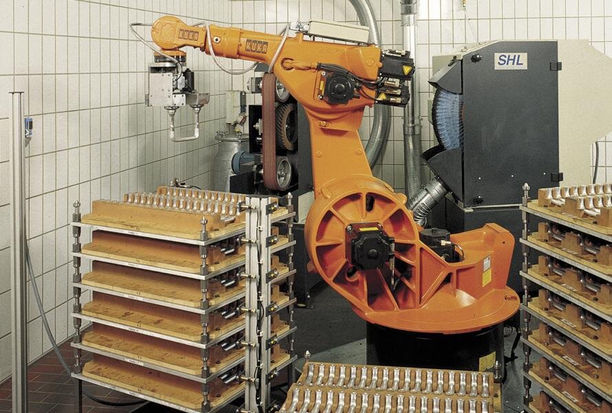 Oranger KUKA Roboterarm bearbeitet metallische Werkstücke auf Ablagen in einer Werkhalle mit weißen Kacheln und SHL-Absauganlage