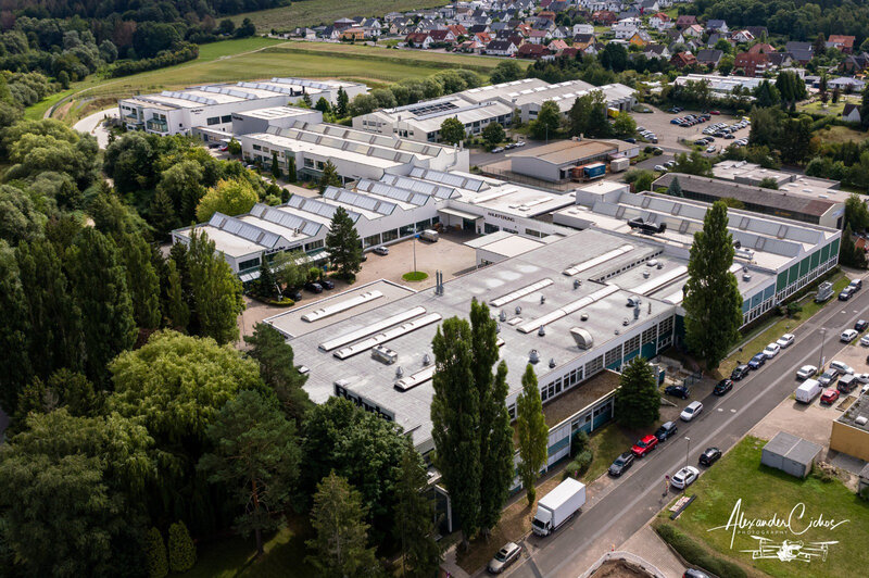 Luftaufnahme eines Industriegebäudes mit mehreren Gebäudeteilen, umgeben von Bäumen und angrenzend an ein Wohngebiet mit vielen Einfamilienhäusern.