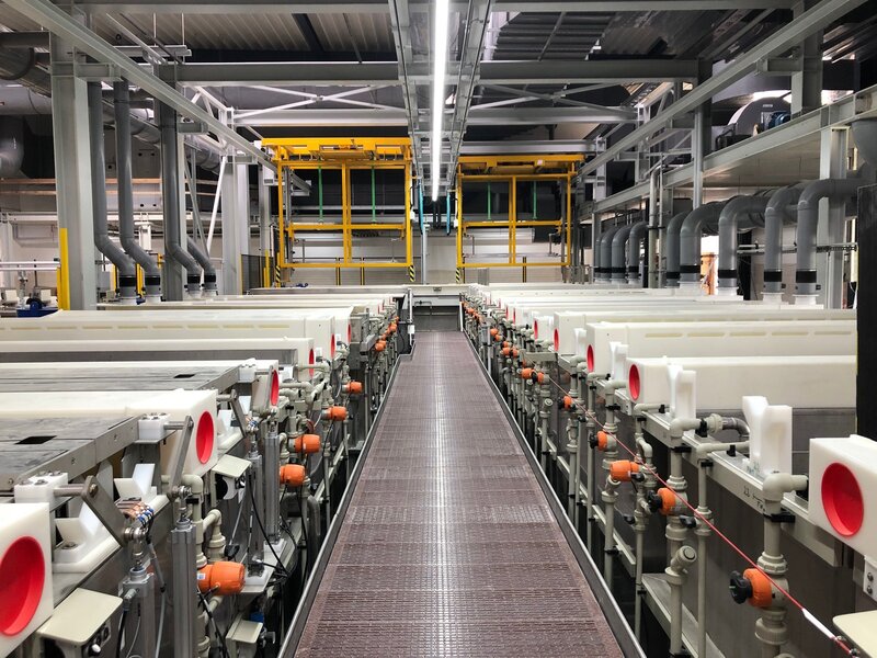 Blick auf eine industrielle Anlage mit parallel angeordneten Kunststofftanks, verbunden mit zahlreichen Rohrleitungen, Ventilen und orangefarbenen Stellmotoren in einer hell beleuchteten Produktionshalle