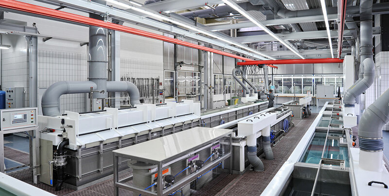 Innenansicht einer sauberen industriellen Produktionsanlage mit großen Wannen, Rohrleitungen und einer arbeitenden Person im Hintergrund