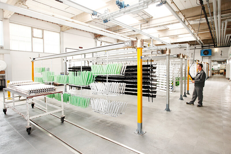 Mitarbeiter bedient eine Trocknungsanlage mit aufgehängten Bauteilen in Grün, Schwarz und Weiß in einer hellen Industrihalle
