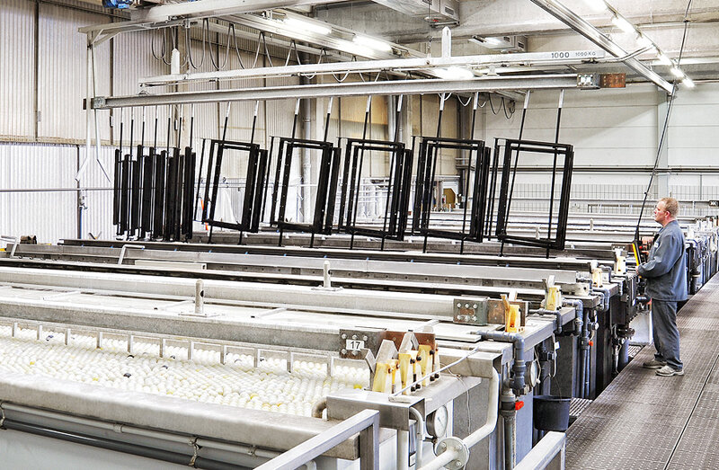 Industrielle Fertigungshalle mit hängenden schwarzen Metallrahmen über mit weißen Kugeln gefüllten Wannen und einem Arbeiter an einer Steuerung