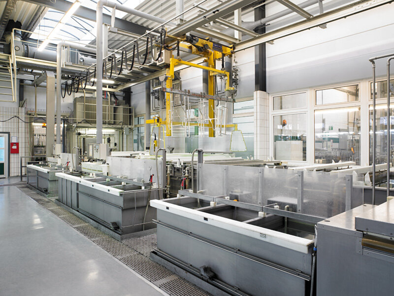 Industrielle Fertigungshalle mit mehreren großen Metallbecken und gelber Hebevorrichtung an Fahrschiene über den Becken