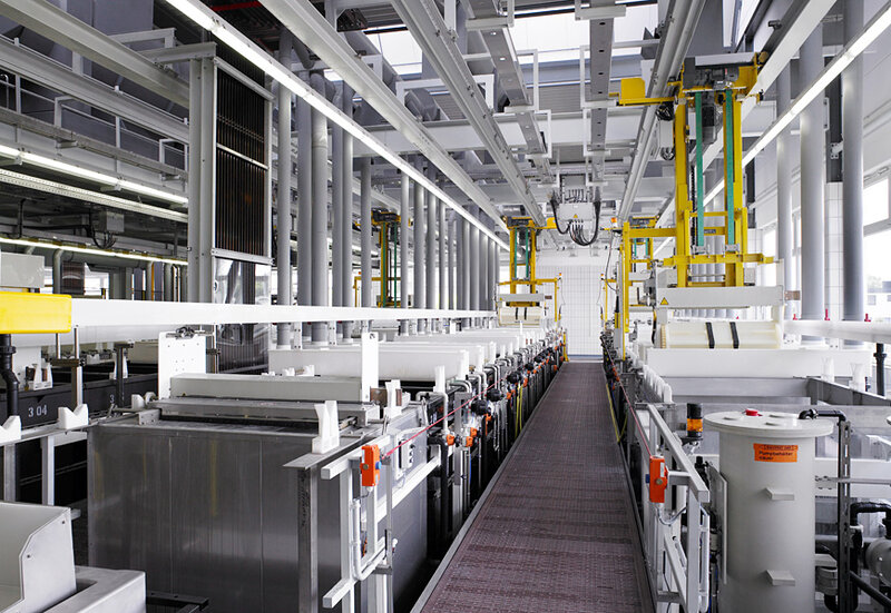 Innenansicht einer industriellen Produktionsanlage mit mehreren rechteckigen Tanks, Hebevorrichtungen und einem erhöhten Steg in der Mitte