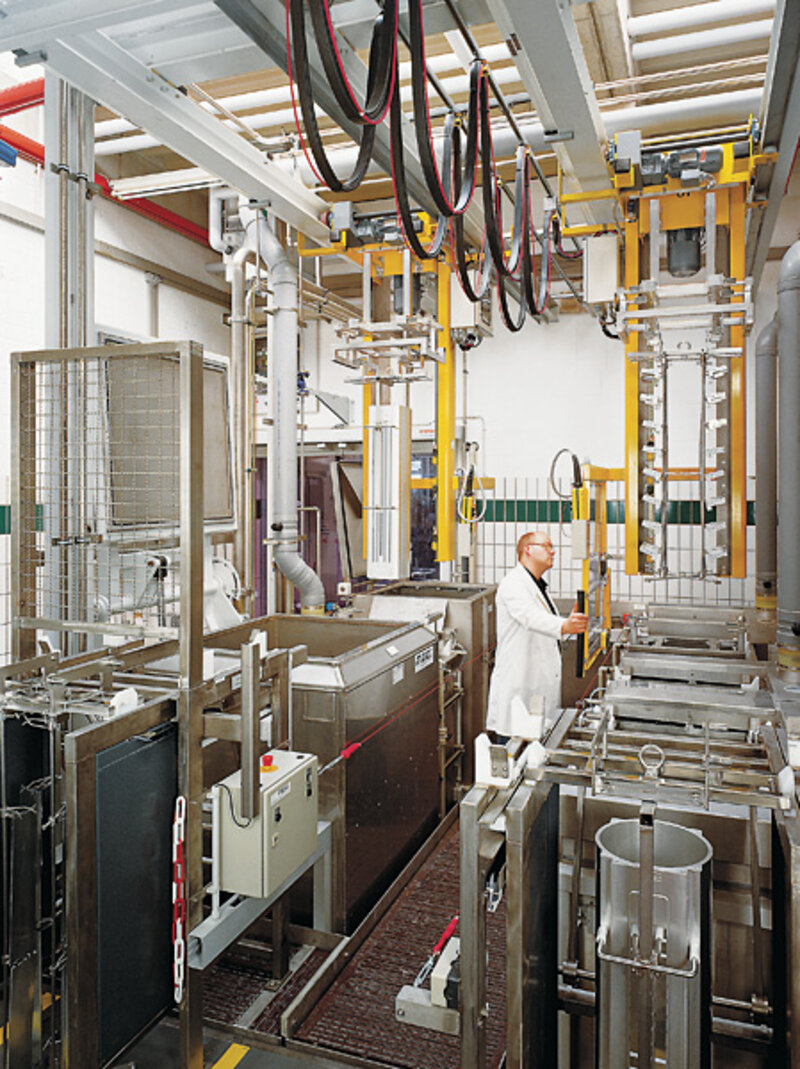 Mann in weißem Kittel bedient eine industrielle Anlage aus metallischen Behältern und Hebevorrichtungen in einer Industriehalle