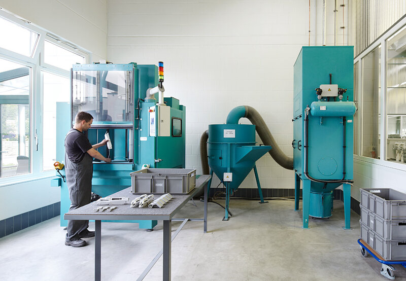 Mann in Arbeitskleidung prüft Werkstück vor Industriemaschine in einem hellen Fabrikraum mit weiteren Maschinen im Hintergrund
