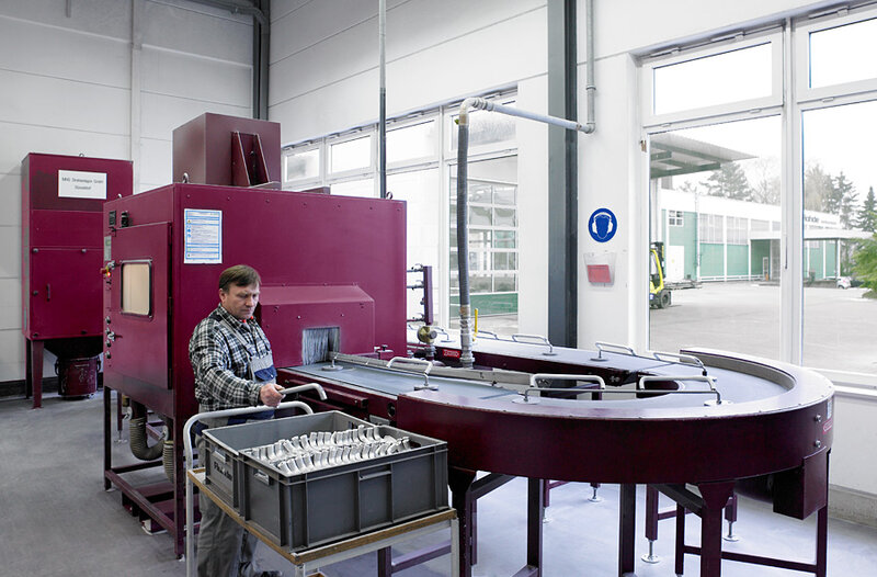 Mann bedient eine große rote Industriemaschine mit gebogenem Förderband in einer hellen Fabrikhalle, vor ihm ein Behälter mit Metallteilen.