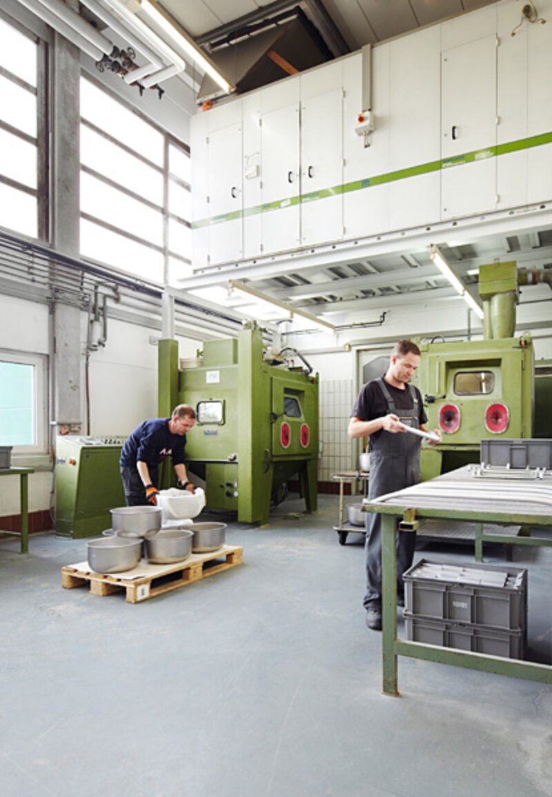 Zwei männliche Arbeiter in einer hellen Produktionshalle bedienen Maschinen und inspizieren Werkstücke, umgeben von grünen Maschinen und Werkzeugen.
