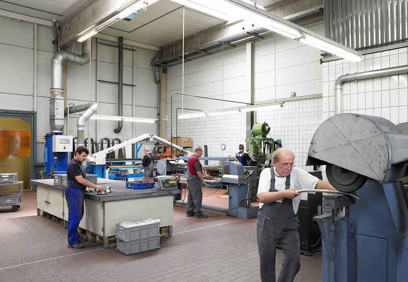 Vier Männer arbeiten in einer hellen industriellen Werkstatt an verschiedenen Maschinen und Werkbänken mit Metallteilen.