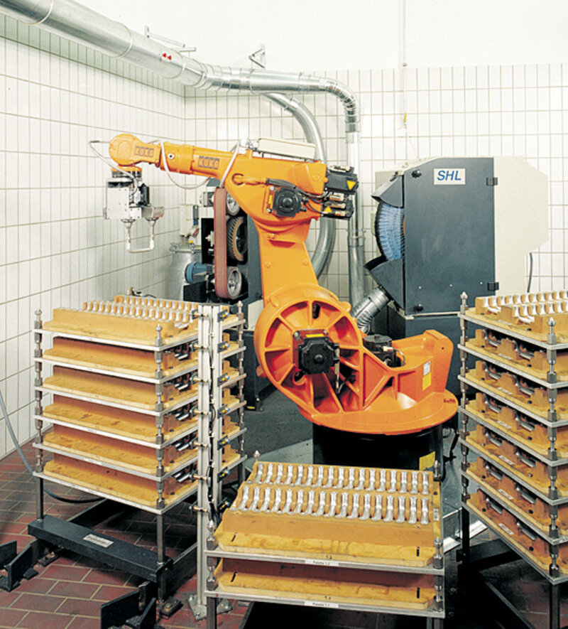 Oranger Industrieroboter KUKA umgeben von mehreren Gestellen mit Metallteilen in einem Raum mit weißen Kachelwänden und Rohrleitungen