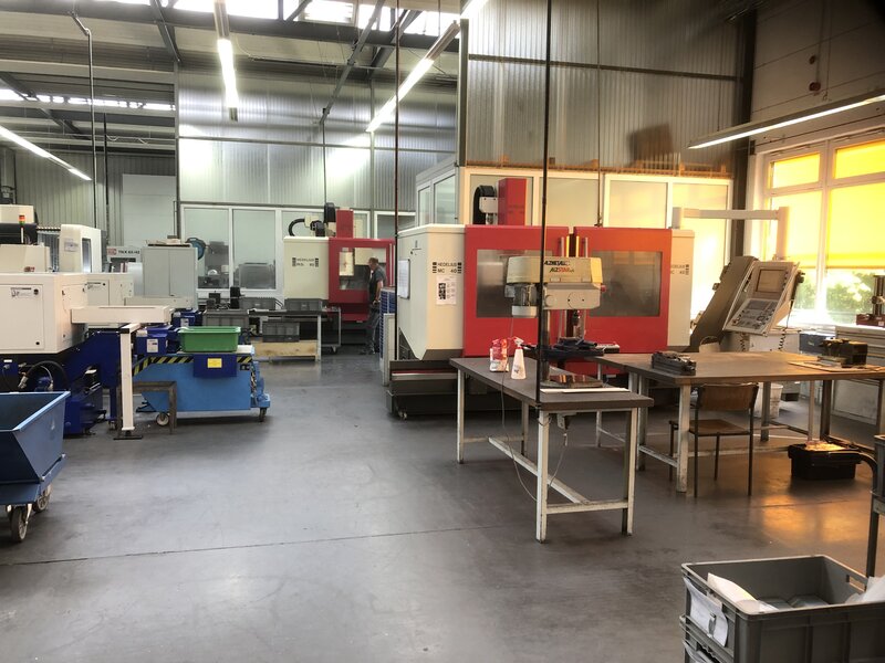 Innenansicht einer Industriehalle mit mehreren CNC-Maschinen, Arbeitstischen, Behältern und einer Person, die eine Maschine bedient.