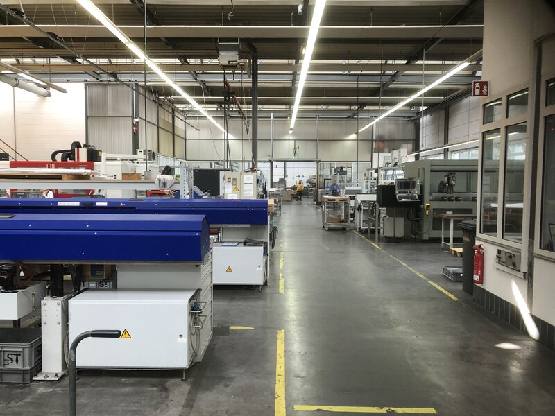Blick in eine Industriehalle mit Maschinenreihen auf der linken Seite, einem breiten Gang in der Mitte und Arbeitsräumen auf der rechten Seite, zwei Personen im Hintergrund