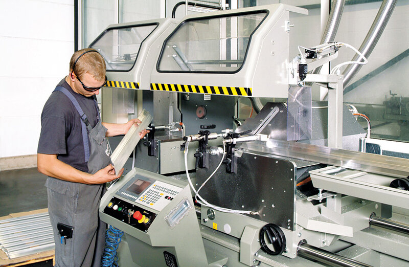 Mann mit Schutzbrille bedient computergesteuerte Metallbearbeitungsmaschine und hält ein bearbeitetes Metallstück in der Hand