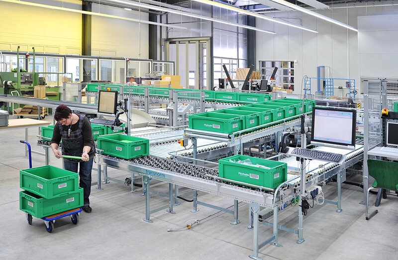 Mann in einem Logistikzentrum neben Förderbändern mit grünen Kunststoffbehältern und Computerarbeitsplatz