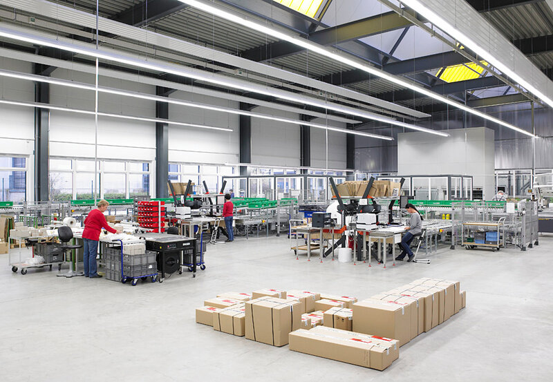 Innenansicht einer Fabrikhalle mit mehreren Arbeitern an Montage- und Verpackungsstationen sowie zahlreichen verpackten Kartons auf dem Boden