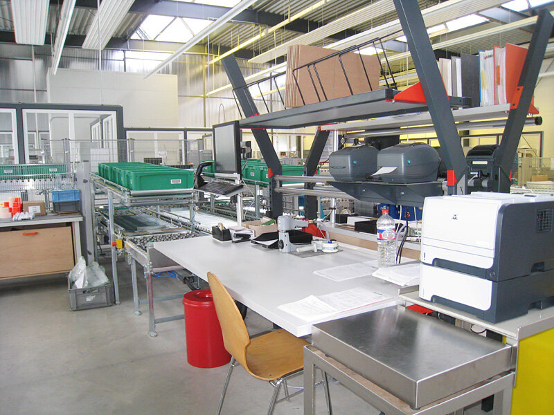 Industrieller Arbeitsplatz mit Tisch, Stuhl, Drucker, Förderband mit grünen Kisten in großer Halle