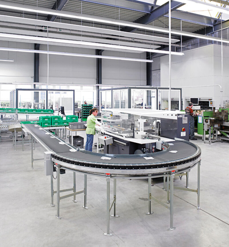 Innenansicht einer Industriehalle mit Förderbandanlage und zwei Mitarbeitern, die an Maschinen arbeiten