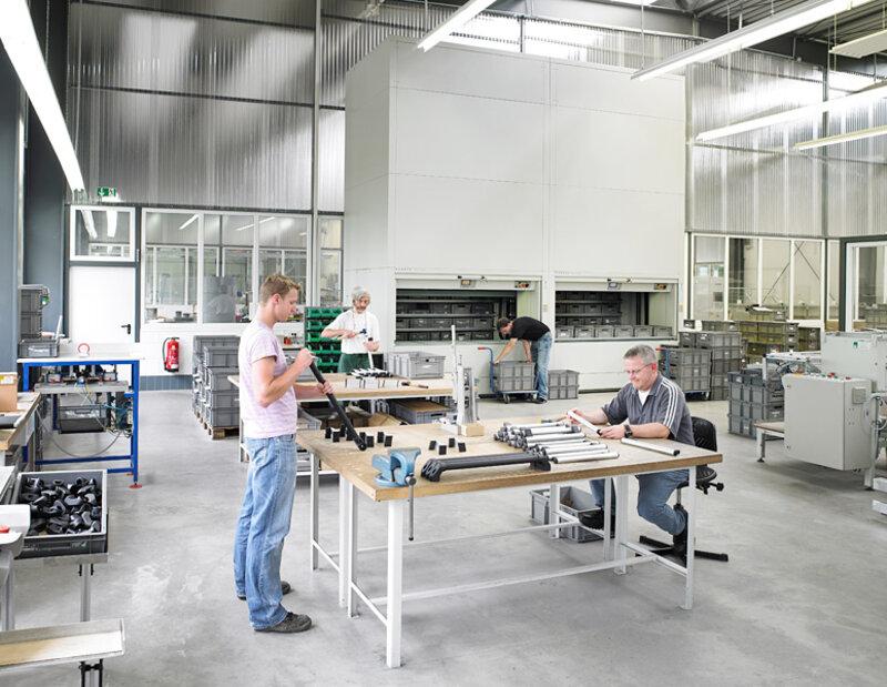 Vier Männer arbeiten an Tischen und Maschinen in einer großen, hellen Fabrikhalle mit Metall- und Kunststoffteilen