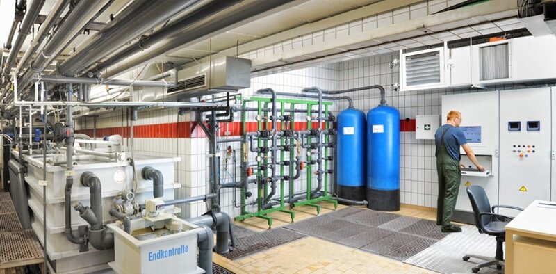 Industrielle Wasseraufbereitungsanlage mit zwei großen blauen Tanks und Rohrleitungen, ein Mitarbeiter bedient ein Steuerpult an der Wand