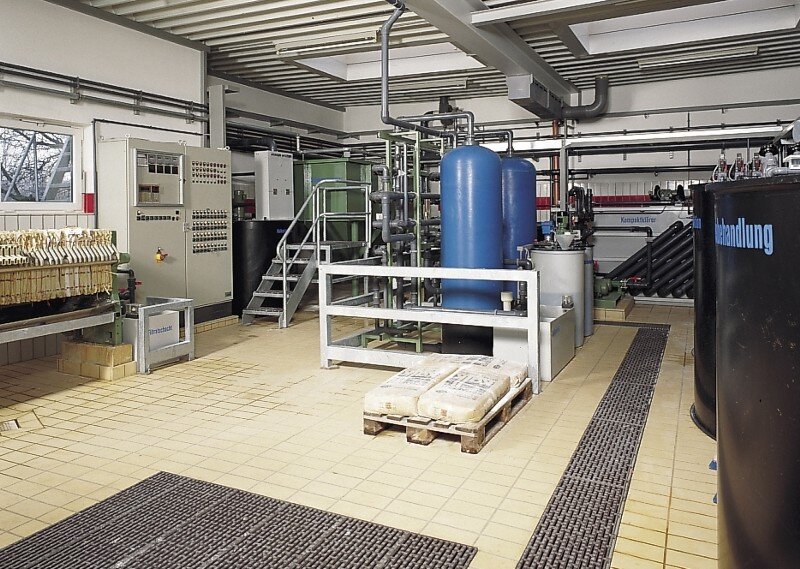Industrieller Raum mit blauen Druckbehältern, Rohrleitungen, Steuerungselementen und einer Holzpalette mit verpackten Säcken auf gefliestem Boden