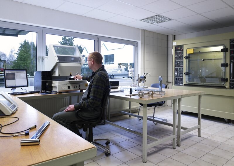 Mann sitzt im Labor an einem Schreibtisch mit einem geöffneten Prüfgerät und einem Computer, im Hintergrund weitere Messgeräte und ein großes industrielles Gerät an der Wand