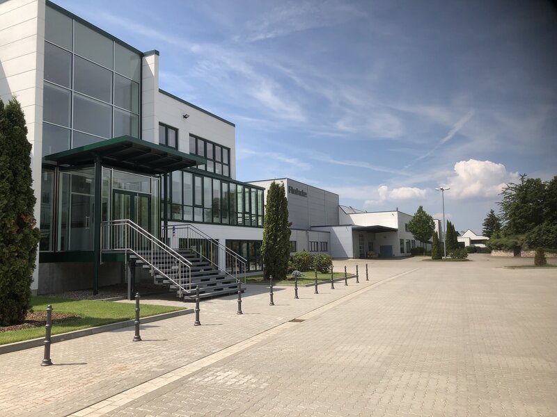 Firmengebäude mit weißer Fassade, großen Fenstern, einem Eingangsbereich mit Treppe und Glashalle, umgeben von Bäumen und gepflasterter Fläche unter bewölktem Himmel