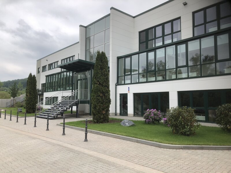 Modernes Bürogebäude mit weißen Wänden und großen Fenstern, davor zwei Nadelbäume und violett blühende Sträucher, gepflasterter Weg mit Metallpfosten davor